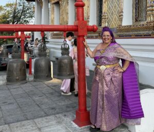 אישה מחייכת לבושה בשמלה תאילנדית מסורתית בצבעי סגול וזהב עומדת ליד פעמונים טקסיים גדולים על מסגרות אדומות מחוץ למקדש מעוטר, כשברקע אנשים אחרים לבושים בבגדים דומים ופרטי מקדש מעוטרים.