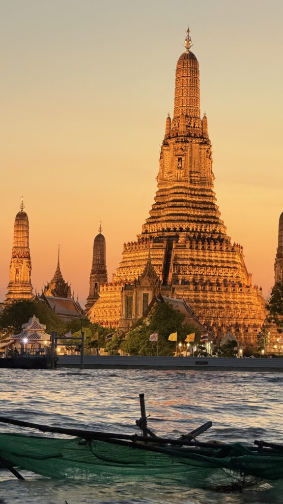 מקדש זהוב עם צריחים מורכבים, וואט ארון, זוהר בשקיעה לצד נהר בבנגקוק, תאילנד. השמיים כתומים, ובחזית נראית סירה במים.