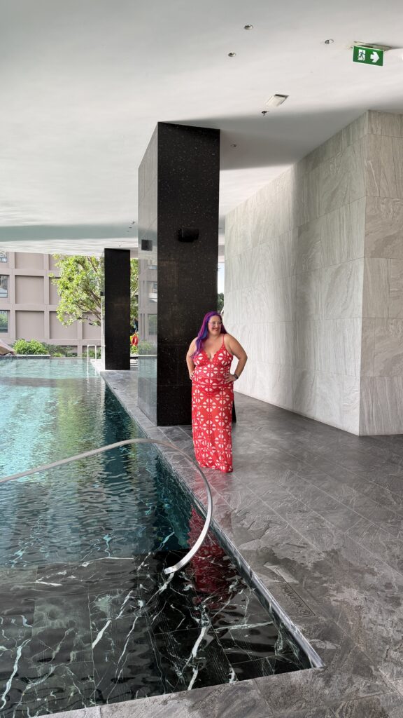 אדם לבוש בשמלה אדומה עם הדפס פרחוני עומד ליד בריכה מקורה עם ריצוף וקירות אפורים. יש אור טבעי, עץ ירוק בחוץ ושלט יציאת חירום על התקרה.