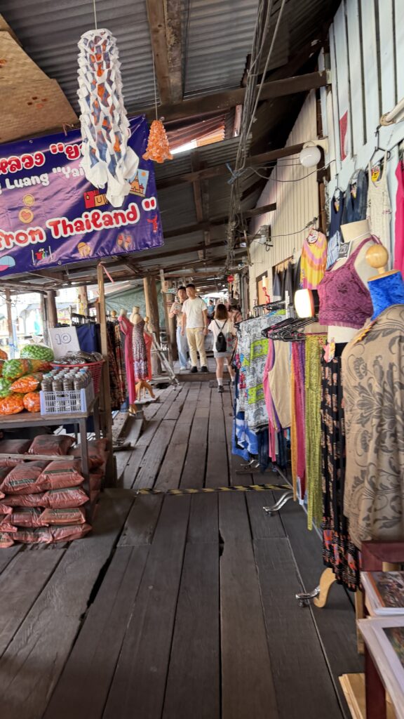 שביל עץ בשוק, עמוס בבגדים צבעוניים, בובות תצוגה ומוצרים מקומיים; שלט "מתאילנד" תלוי מעל, וברקע נראים אנשים המסתכלים על המוצרים.