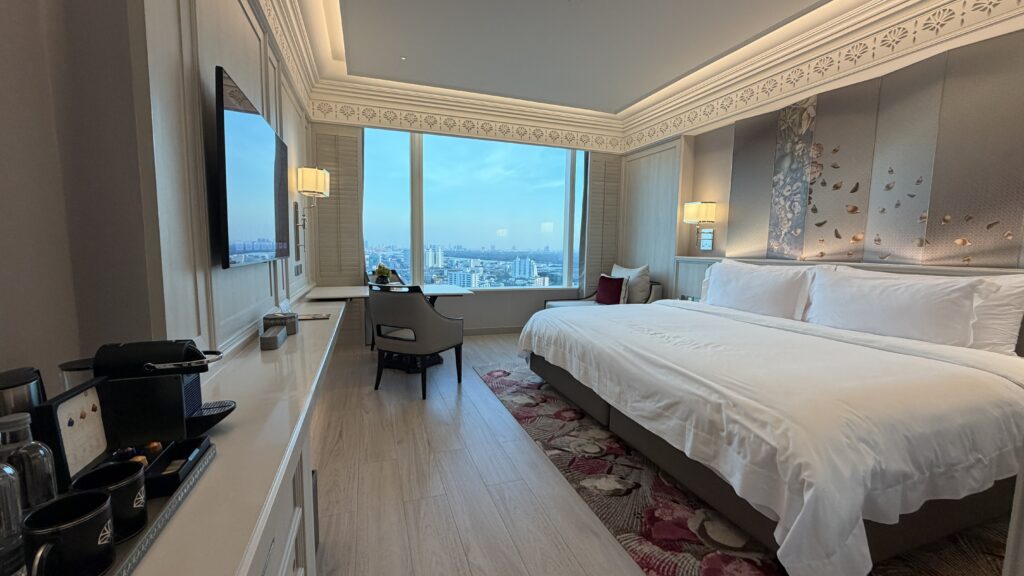 חדר מלון מפואר עם מיטה גדולה, טלוויזיה המותקנת על הקיר, שולחן ליד חלון גדול המשקיף על נוף העיר, מכונת קפה ועיצוב אלגנטי עם תאורה רכה.