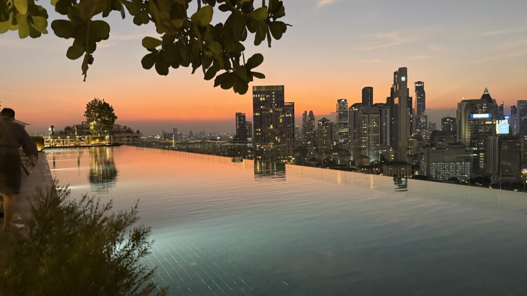 בריכת אינסוף משקיפה על קו הרקיע של העיר בשעת השקיעה, עם שמיים צבעוניים ובניינים גבוהים המשתקפים במים. עלים וצמחים ממסגרים את הנוף, בעוד אדם הולך לאורך קצה הבריכה משמאל.