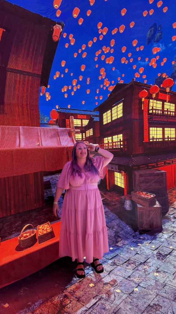 אישה בשמלה ורודה עומדת ברחוב מרוצף אבן, בין בניינים מוארים בפנסים מעץ ופנסים זוהרים מרחפים בשמי הלילה, ויוצרים אווירה חגיגית.