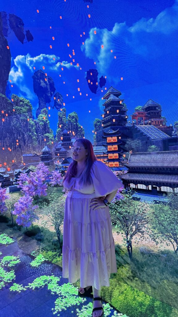 אישה בשמלה סגולה בהירה עומדת מול רקע דיגיטלי מרהיב וצבעוני, המציג פנסים, פריחת דובדבן, הרים וארכיטקטורה מסורתית של מזרח אסיה, הזוהרים תחת שמיים כחולים.