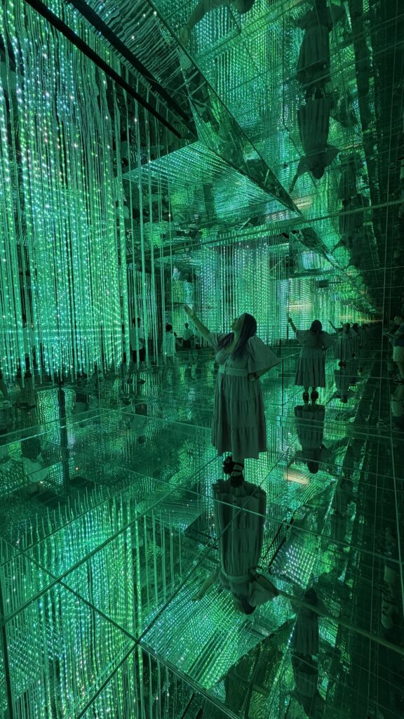 אדם לבוש שמלה לבנה עומד בחדר מרוצף מראות, המלא בשרשראות נורות LED ירוקות תלויות, היוצרות השתקפויות אינסופיות ואווירה עתידנית ומרתקת.