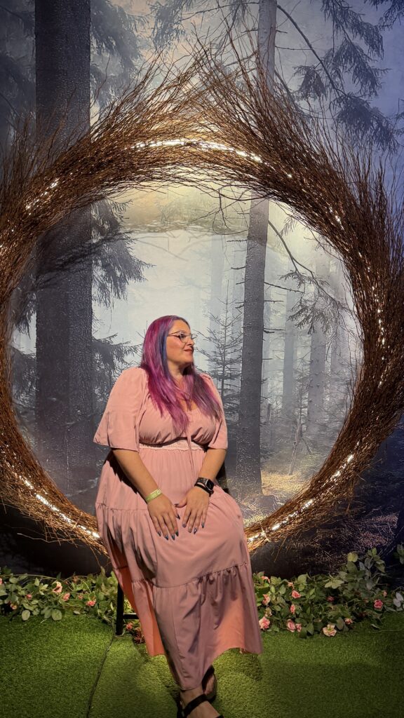 אישה עם שיער ארוך ורוד וסגול, המרכיבה משקפיים ולובשת שמלה ורודה, יושבת על כיסא בתוך זר עגול גדול עם אורות, על רקע יער ערפילי. פרחים וצמחייה מקשטים את הקרקע.