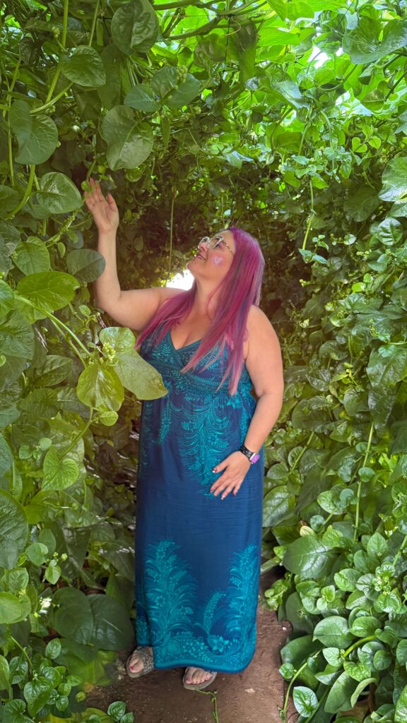 אישה עם שיער ורוד ארוך בשמלה כחולה עומדת מוקפת בגפנים ירוקות ושופעות, מושיטה את ידה כדי לגעת בעלים ומחייכת כשהיא מביטה בצמחייה מעליה.