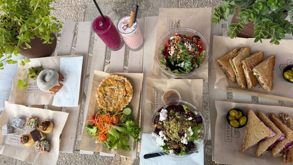 שולחן ערוך במגוון מנות, כולל כריכים, סלטים, קיש, מאפינס, עוגות, שני משקאות פירותיים ועציצים קטנים לקישוט. האוכל נראה טרי וצבעוני, מסודר על נייר חום וצלחות לבנות.