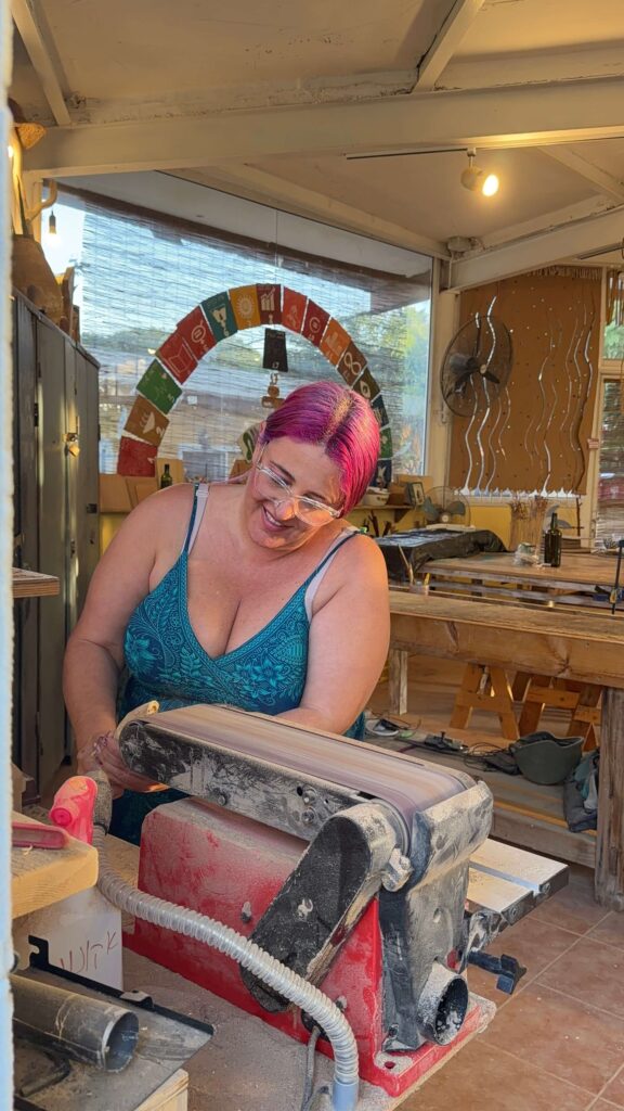 אישה עם שיער ורוד ומשקפיים, מחייכת בזמן שהיא משתמשת במכונת ליטוש על פיסת עץ בסדנה מוארת היטב, מלאה בכלים ובשולחנות עבודה מעץ.