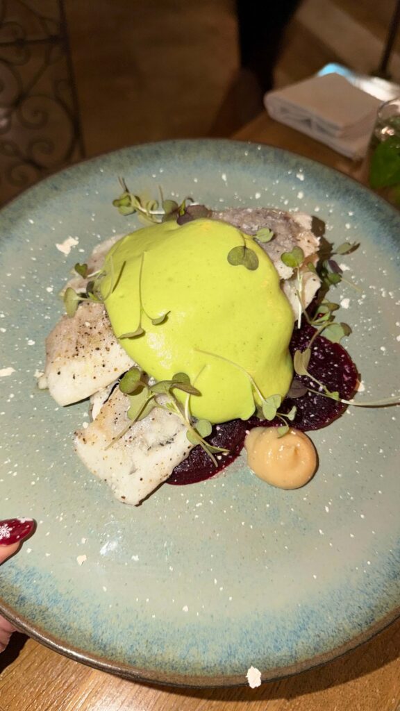 מנה המורכבת מפילטים של דג לבן עם קצף ירוק, מוגשת עם פרוסות סלק, נבטים קטנים וכמות קטנה של רוטב בז' על צלחת קרמיקה כחולה.