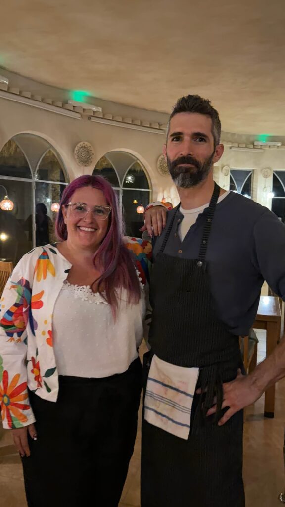 אישה עם שיער סגול ומשקפיים עומדת מחייכת ליד גבר מזוקן בחלוק כהה בתוך מסעדה מוארת באור חם עם חלונות מקושתים ומנורות עגולות תלויות.