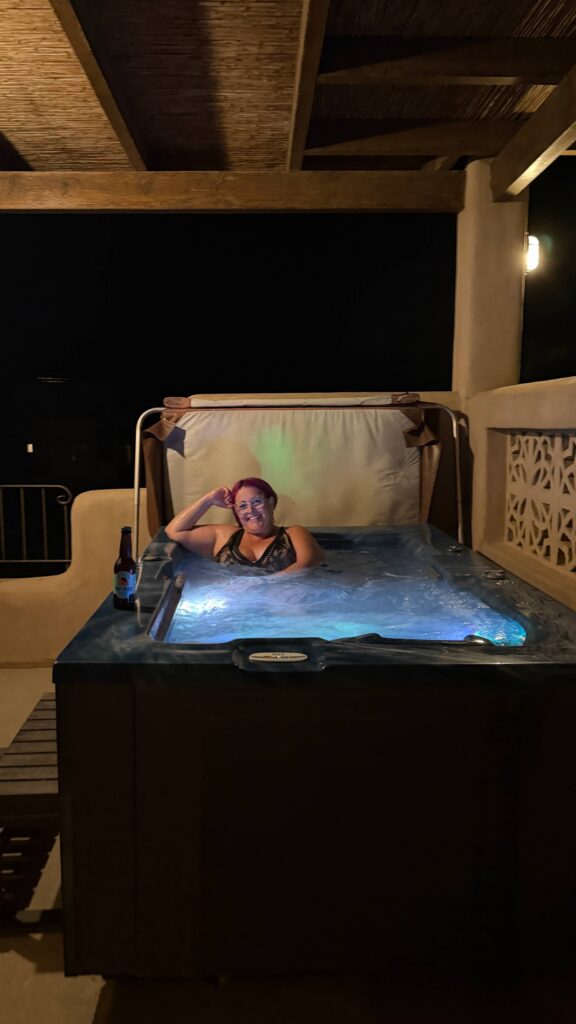 אדם עם שיער ורוד נרגע בג'קוזי חיצוני בלילה, מחייך ונשען על הקצה. ליד הג'קוזי מונחת בקבוק בירה, והפטיו מואר בתאורה רכה.