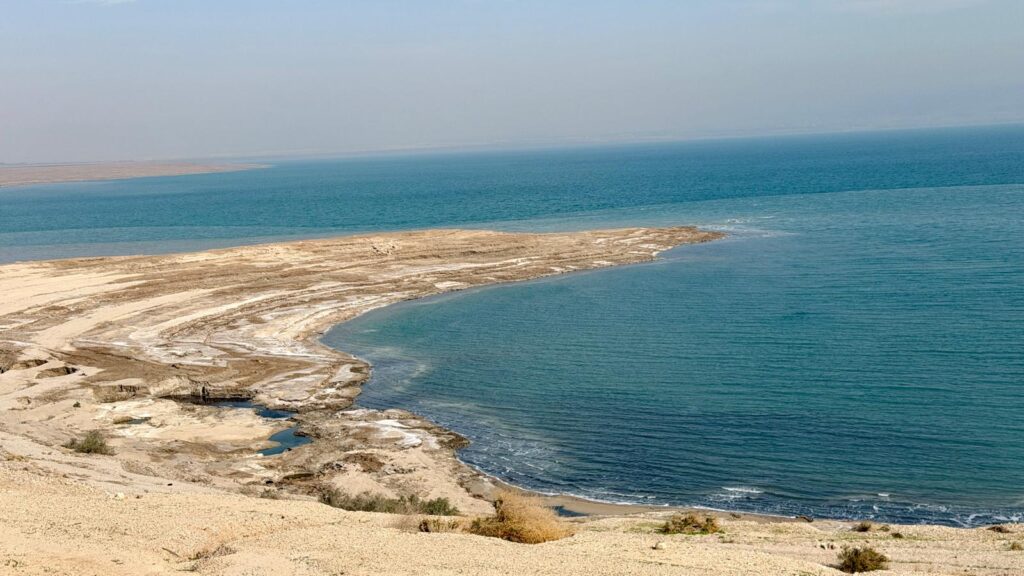 נוף רחב של קו החוף של ים המלח, המציג שכבות של מלח ומינרלים על החוף החולי לצד המים הכחולים והשקטים תחת שמים מעורפלים.