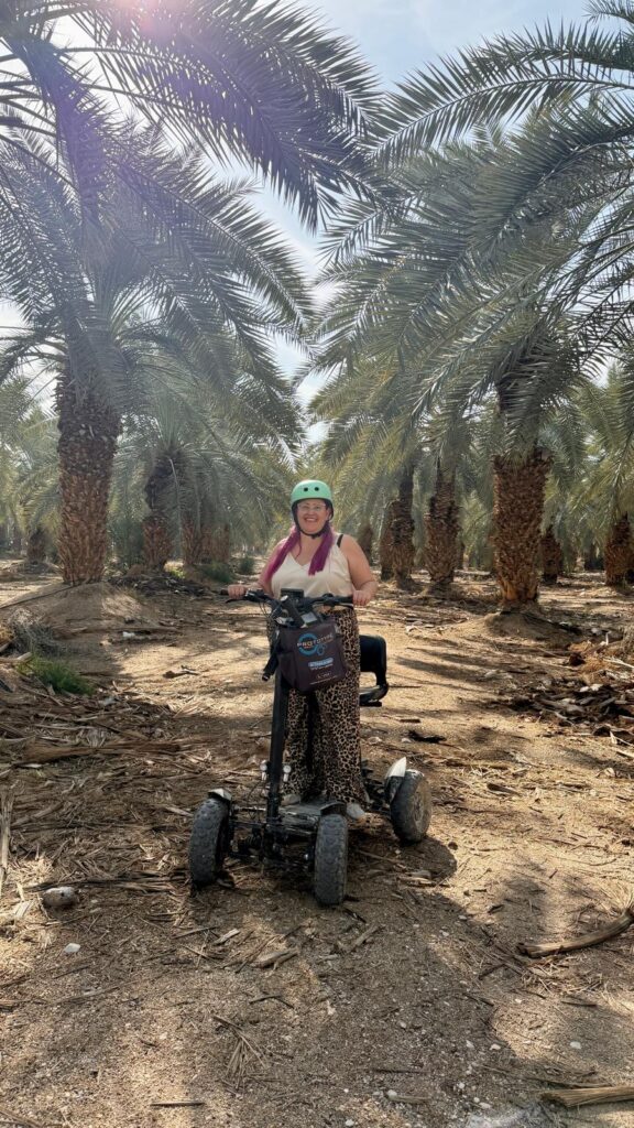 אישה חובשת קסדה ירוקה ומכנסיים עם הדפס חיות מחייכת כשהיא יושבת על קטנוע ניידות שטח בין עצי דקל מוארים בשמש.