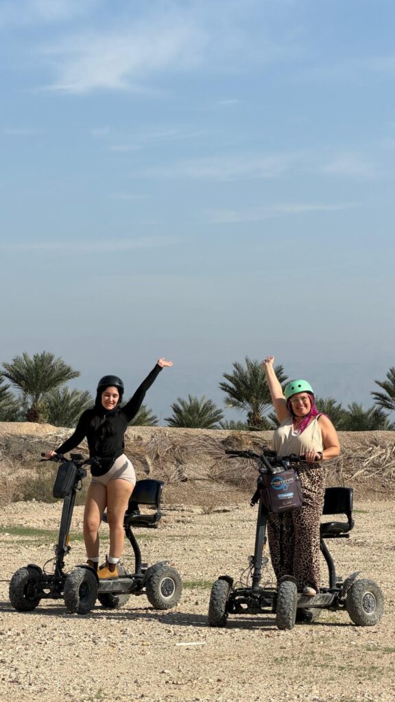 שתי נשים חובשות קסדות ולבושות בבגדים קז'ואליים רוכבות על קטנועים חשמליים שטח על משטח סלעי, מחייכות ומרימות זרוע אחת באוויר. ברקע נראים עצי דקל ושמיים כחולים.