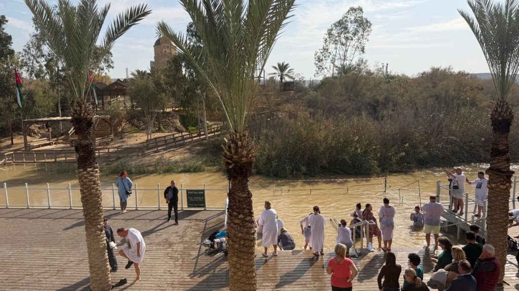 אנשים לבושים בגלימות לבנות מתאספים על סיפון עץ ליד נהר בוצי, מוקפים בעצי דקל וקנים, כשברקע נראים כנסייה ודגלים תחת שמים מעוננים חלקית.