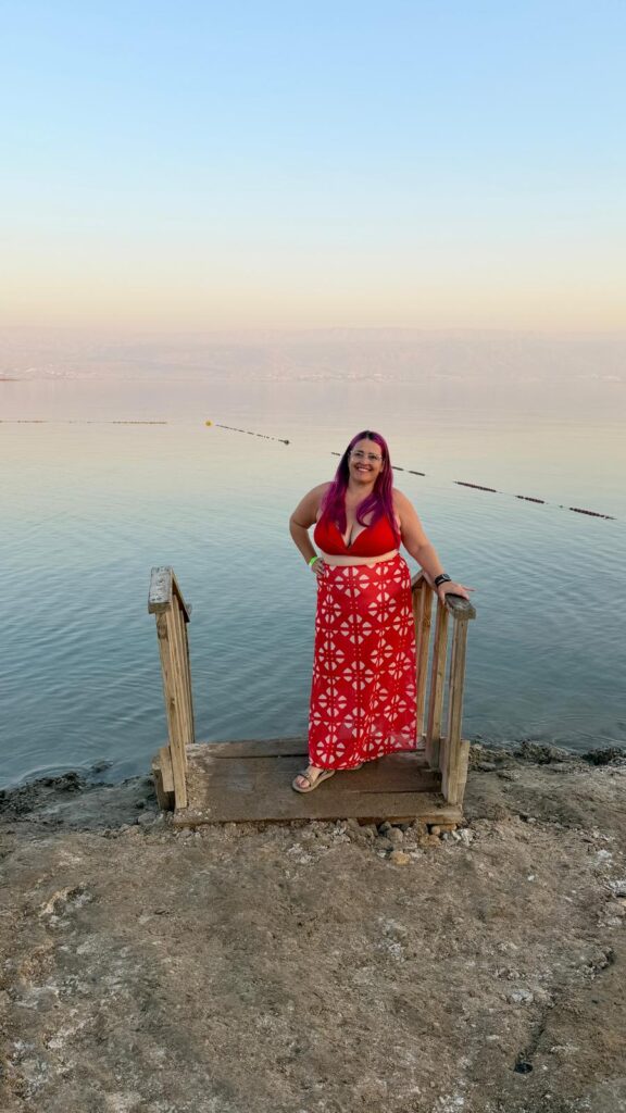 אישה עם שיער ורוד ארוך, לבושה בשמלה אדומה-לבנה, עומדת מחייכת על גרם מדרגות עץ המוביל אל מים רגועים ורחבים תחת שמיים בהירים.