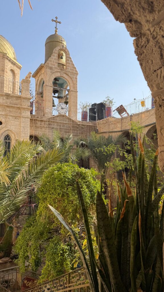 חצר כנסייה מאבן עם צמחייה ירוקה ושופעת, בה מתנשא מגדל פעמונים עם צלב ושעון, תחת שמיים כחולים וצלולים. הארכיטקטורה מעוטרת, עם קשתות וכיפות בולטות לעין.