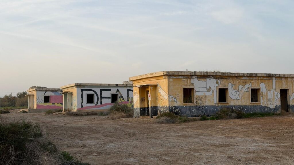 מבנים נטושים מבטון צהוב ומכוסים גרפיטי ניצבים על אדמה צחיחה ויבשה תחת שמים מעוננים. שיחים וצמחייה דלילה נראים בקדמת התמונה וברקע.