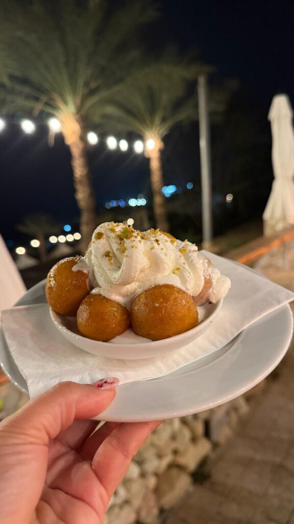 יד אוחזת בצלחת עם גולאב ג'מון ומעליה שכבה נדיבה של קצפת, מקושטת באגוזים קצוצים. עצי דקל ונורות מחרוזת מקשטים את התפאורה הלילית החיצונית ברקע.