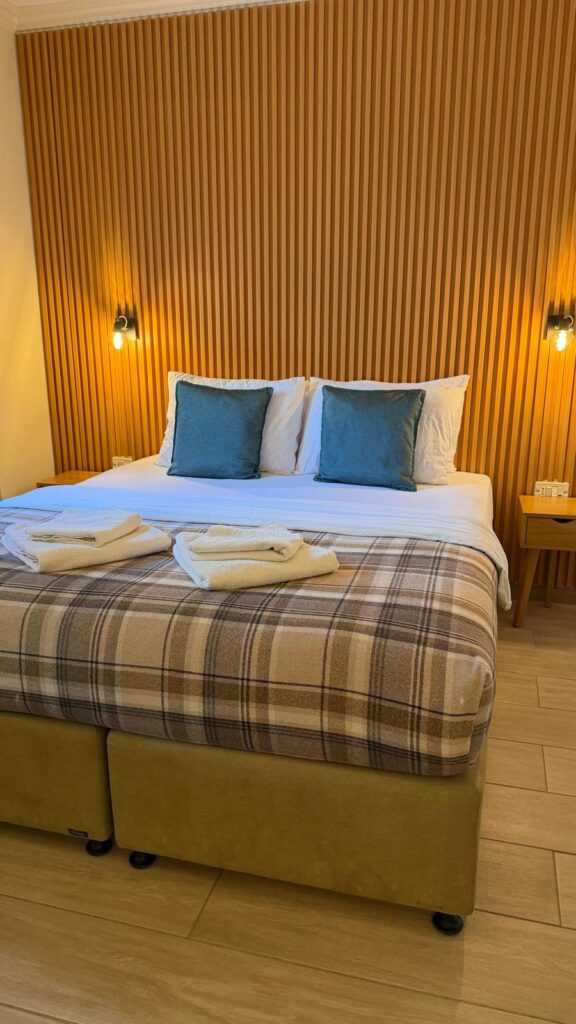 מיטה מסודרת עם כריות נוי כחולות, שמיכה משובצת ושתי מגבות מקופלות מונחת על רקע קיר עץ עם שני פמוטים קיר, בחדר שינה נעים ומודרני.