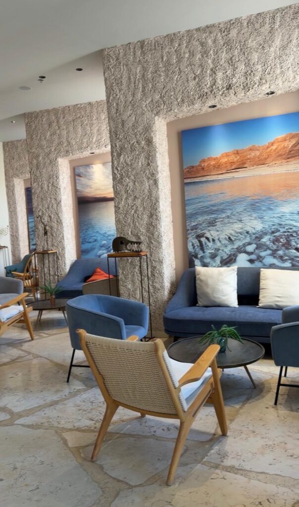 אזור ישיבה מודרני עם ספות כחולות, כיסאות עץ, שולחנות קטנים ותמונות קיר של נופים הכוללים מים והרים. החלל כולל קירות מרקמים ורצפת אבן בהירה, היוצרים אווירה נינוחה ומזמינה.