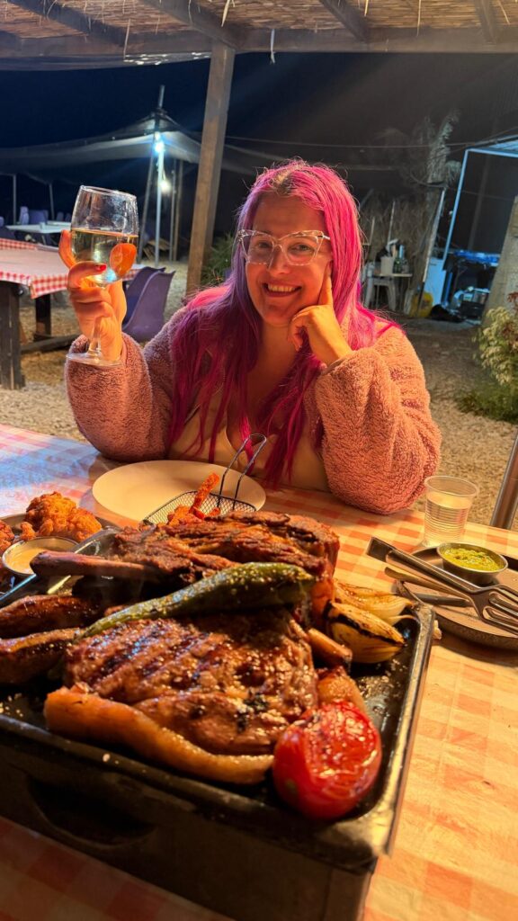 אישה מחייכת עם שיער ורוד ארוך ומשקפיים מרימה כוס יין לבן בשולחן בחוץ, כשברקע מונחת מגש גדול עם בשרים וירקות בגריל. התאורה חמימה והאווירה נינוחה.