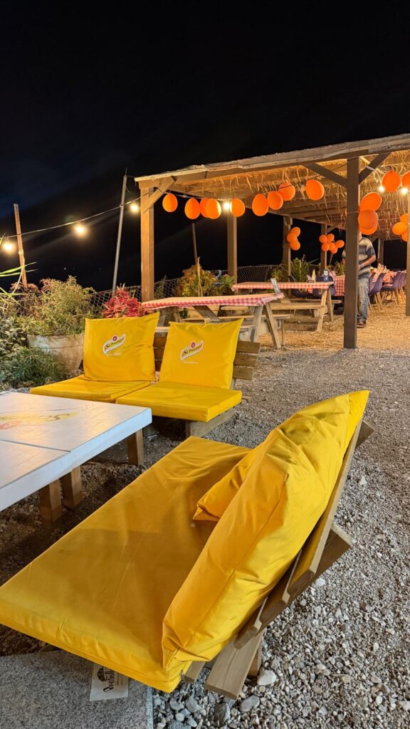 אזור ישיבה חיצוני בלילה עם כיסאות מרופדים בצבע צהוב בוהק, שולחנות עץ ונורות מחרוזת. בלונים כתומים מקשטים את אזור האוכל המקורה עם מפות משובצות. סביב ניתן לראות אדמת חצץ וצמחים.