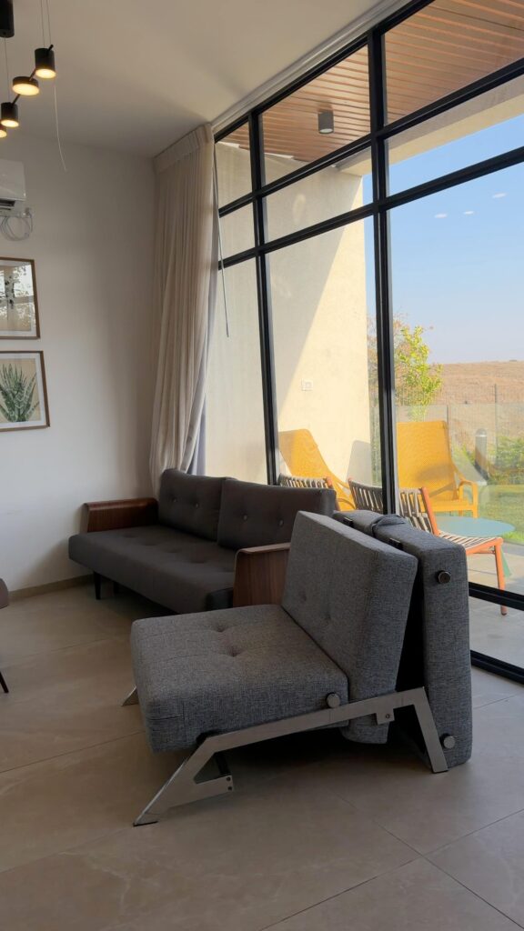 בחזית נראית כורסת ספה נפתחת בצבע אפור, וספה אפורת תואמת המוצבת מול הקיר בסלון מואר באור השמש, עם חלונות מהרצפה עד התקרה.