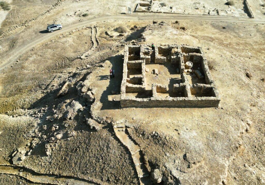 מבט אווירי על חורבות אבן עתיקות בנוף מדברי, עם כמה אנשים העומדים בסמוך ורכב חונה על דרך עפר ברקע.