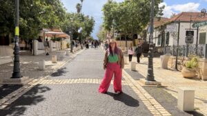 אדם חובש כובע קש ולובש מכנסיים ורודים בוהקים עומד באמצע רחוב מרוצף אבן, המוקף בעצים, בחנויות ובהולכי רגל אחרים.