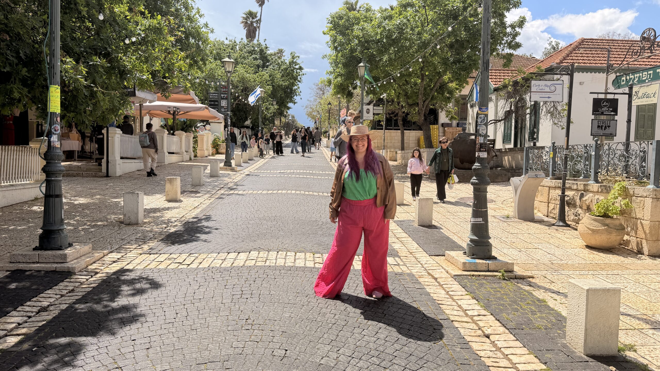 אדם חובש כובע קש ולובש מכנסיים ורודים בוהקים עומד באמצע רחוב מרוצף אבן, המוקף בעצים, בחנויות ובהולכי רגל אחרים.