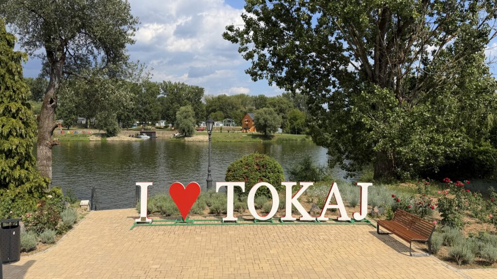 אותיות לבנות גדולות מרכיבות את הכיתוב "I ❤️ TOKAJ" לצד נהר, מוקפות בצמחייה, עצים ופרחים. ספסל ניצב בסמוך, והשמיים מעוננים חלקית עם כתמים כחולים מעל האזור השלווה והציורי.