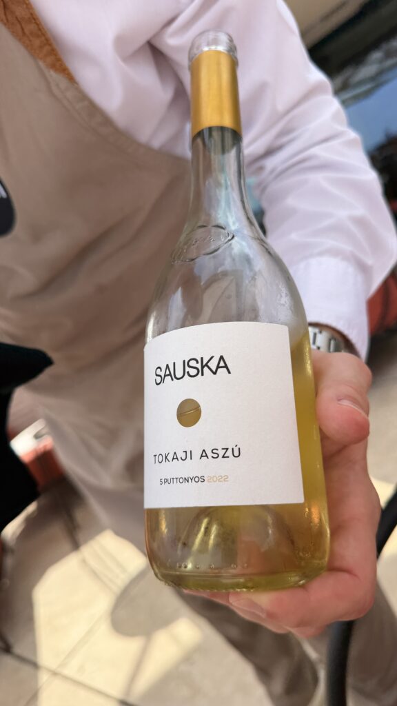 אדם אוחז בבקבוק יין "Sauska Tokaji Aszú 5 Puttonyos 2022", עם תווית לבנה ואלמנטים מוזהבים. האיש לובש חולצה בהירה וסינר בצבע בז'.