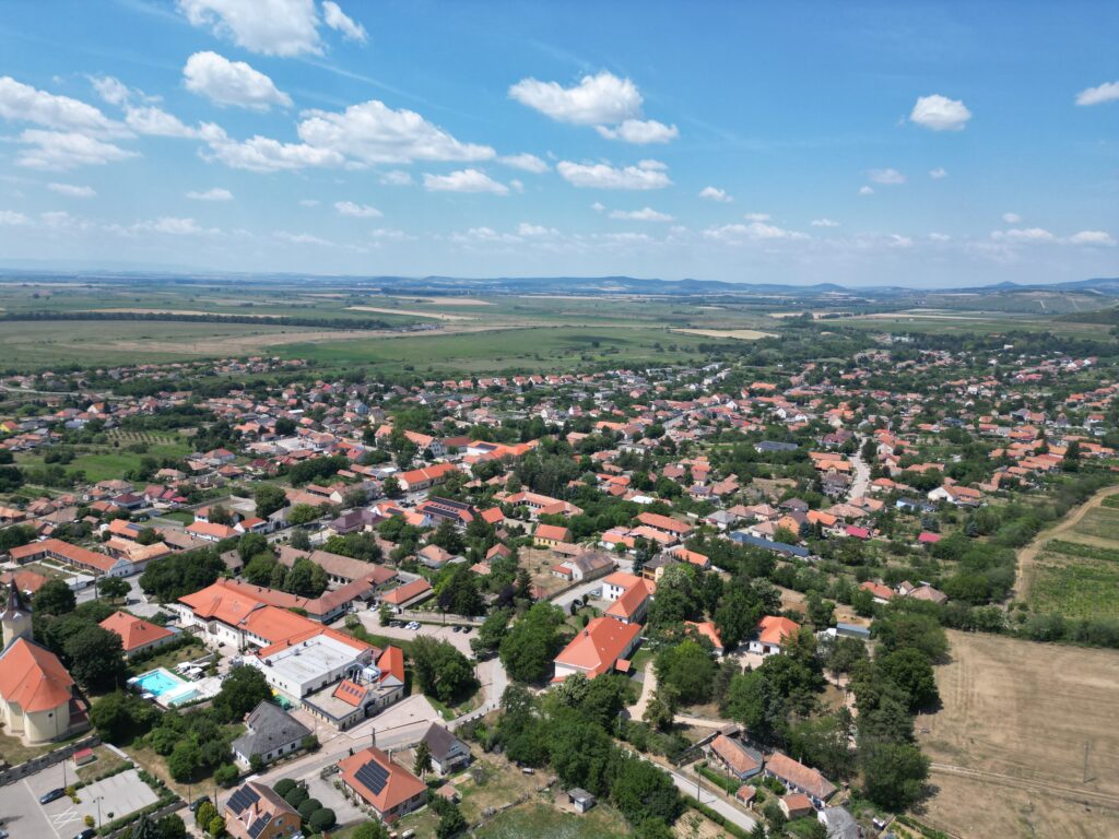 מבט אווירי על עיירה קטנה עם בתים בעלי גגות אדומים, עצים ירוקים ושדות פתוחים תחת שמיים כחולים עם עננים פזורים. גבעות ושטחי חקלאות נפרשים אל האופק.