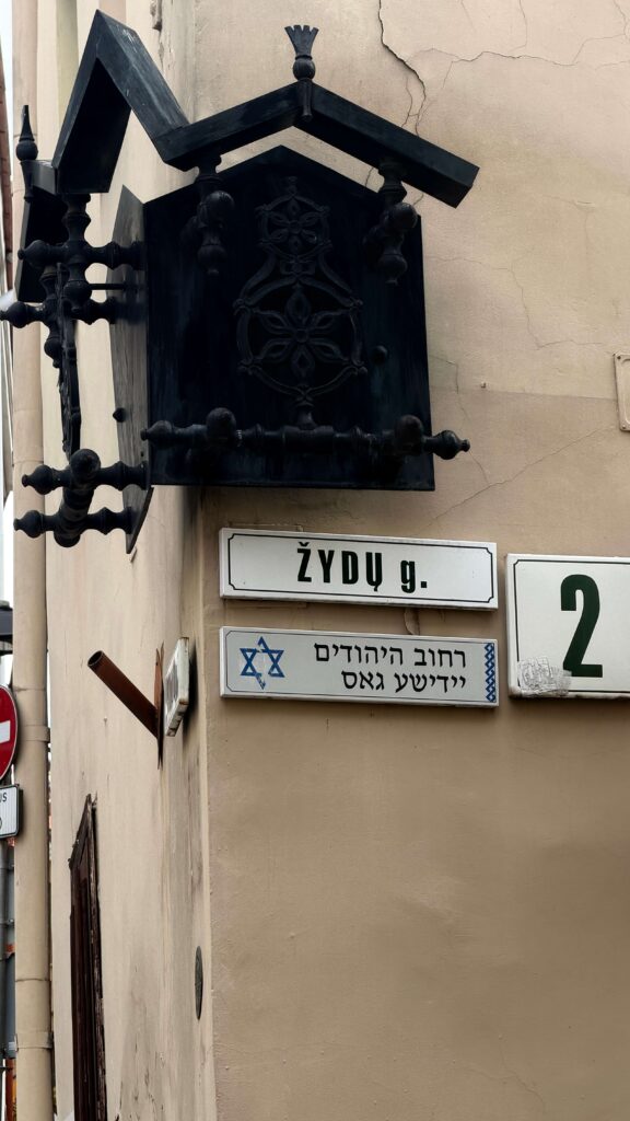 קיר בצבע בז' ועליו פנס שחור דקורטיבי מעל שלטי רחוב, באחד מהם כתוב “Žydų g.” ובאחר טקסט בעברית וביידיש, מגן דוד והמספר 2 על לוחית לבנה.