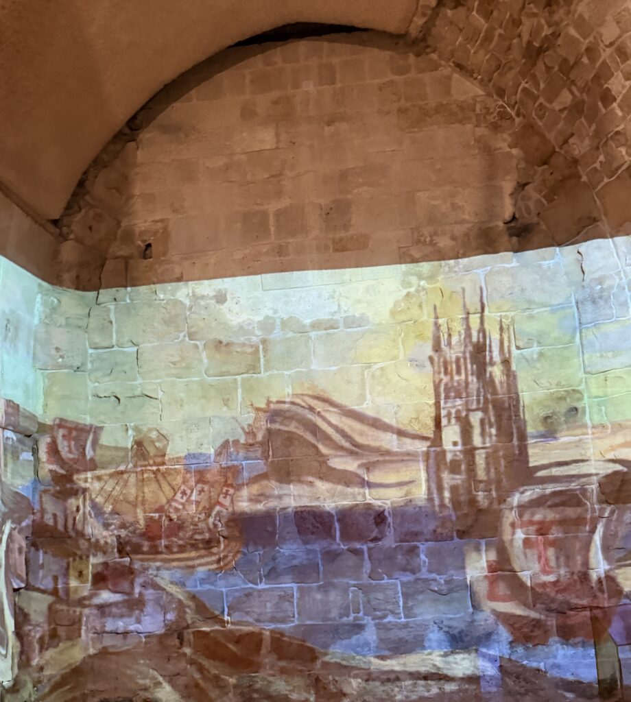 נוף עירוני היסטורי וצבעוני מוקרן על קיר אבן בתוך חדר מקושת, ובו נראים מבנים, קתדרלה ונהר.