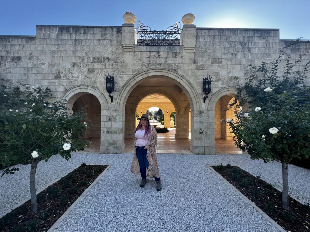 אדם חבוש כובע ולבוש במעיל ארוך עומד על שביל חצץ בין שני שיחי ורדים, מול מבנה אבן בעל שלושה קשתות ופנסים. אור השמש חודר מבעד לקשת המרכזית.
