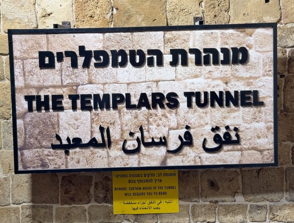 על קיר אבן תלויה שלט שעליו כתוב “מנהרת הטמפלרים” בעברית, באנגלית ובערבית, ומתחתיו מופיעה אזהרה צהובה המציינת כי באזורים מסוימים יש להתכופף.